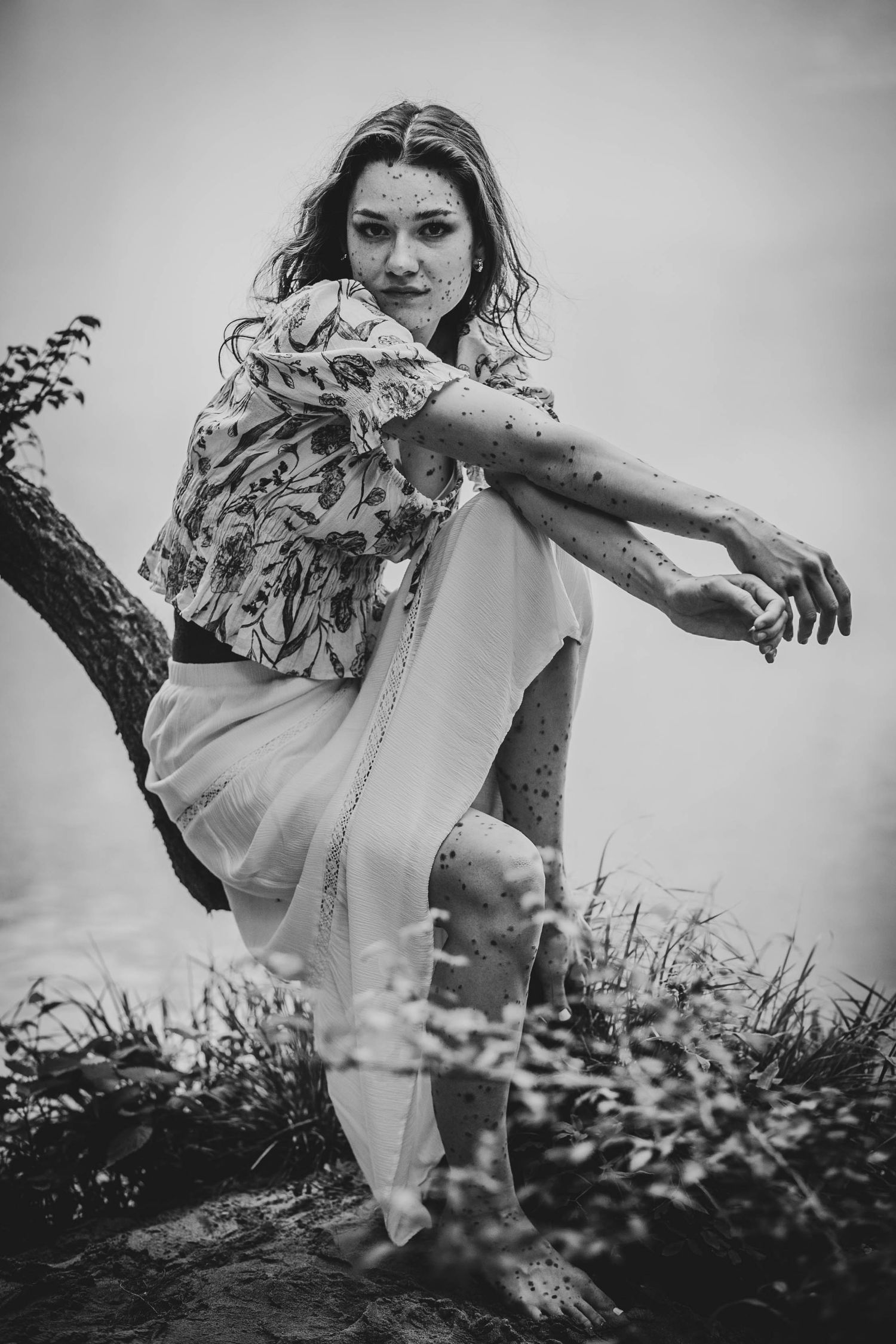 Black and white artistic photo of a woman in floral top and flowy pants balanced on a tree branch in nature.