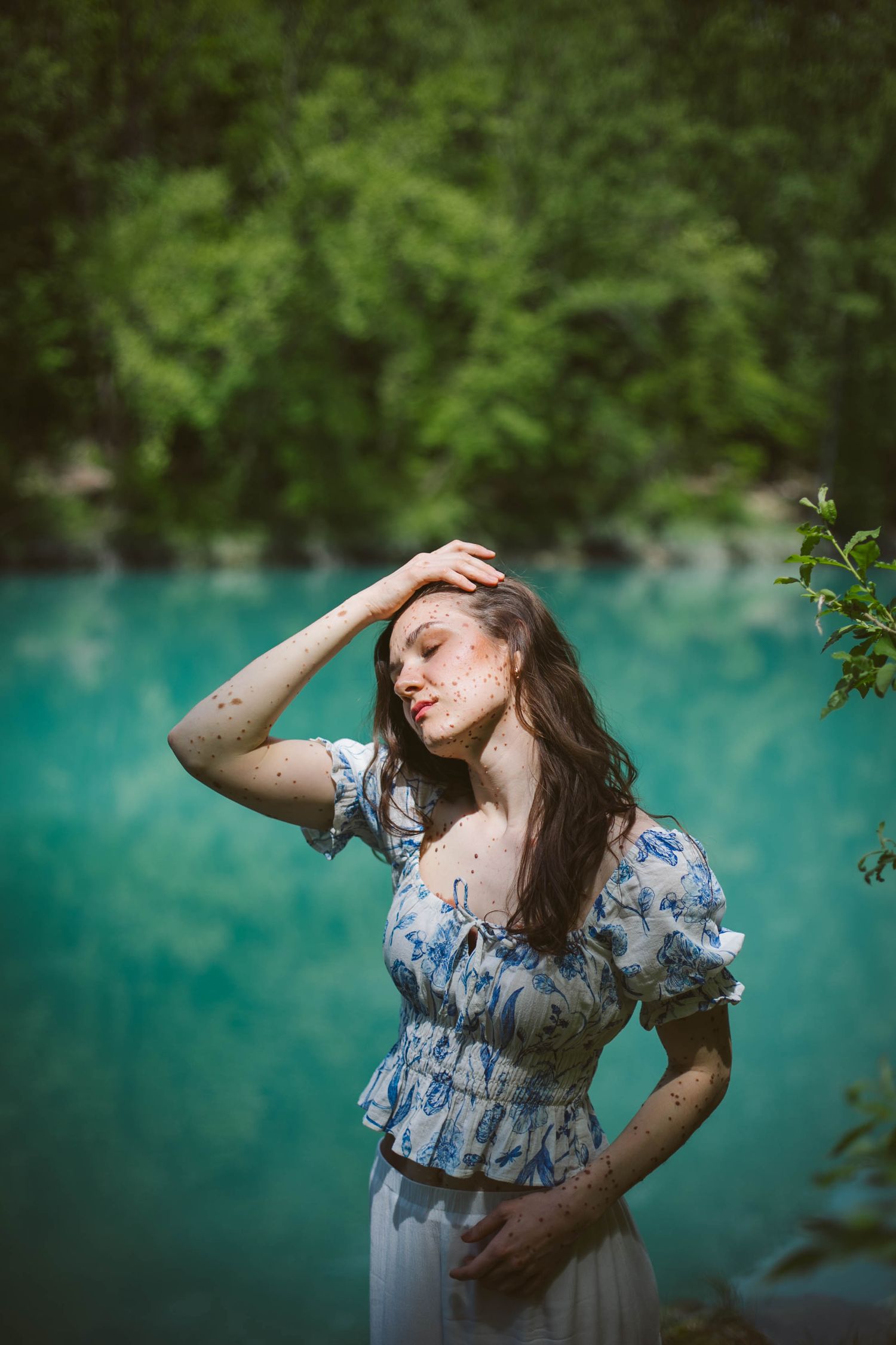 A woman in a flowing blue floral dress poses gracefully near a turquoise lake on a summer day.