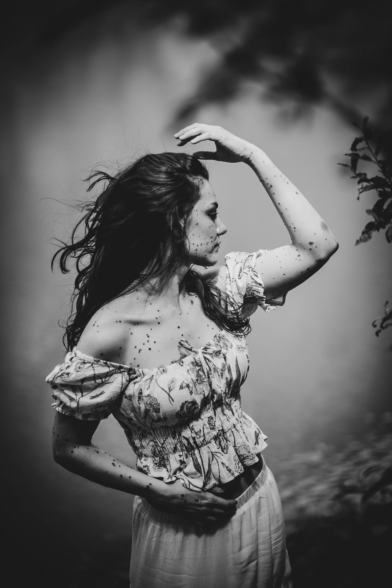 Dramatic black and white portrait of a figure in floral dress striking an emotive pose against stormy clouds.