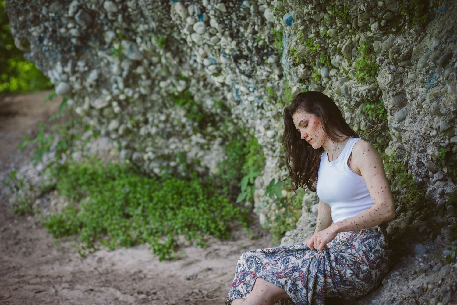 A person in a white top and floral skirt sits pensively against a rocky wall surrounded by blooming spring flowers.