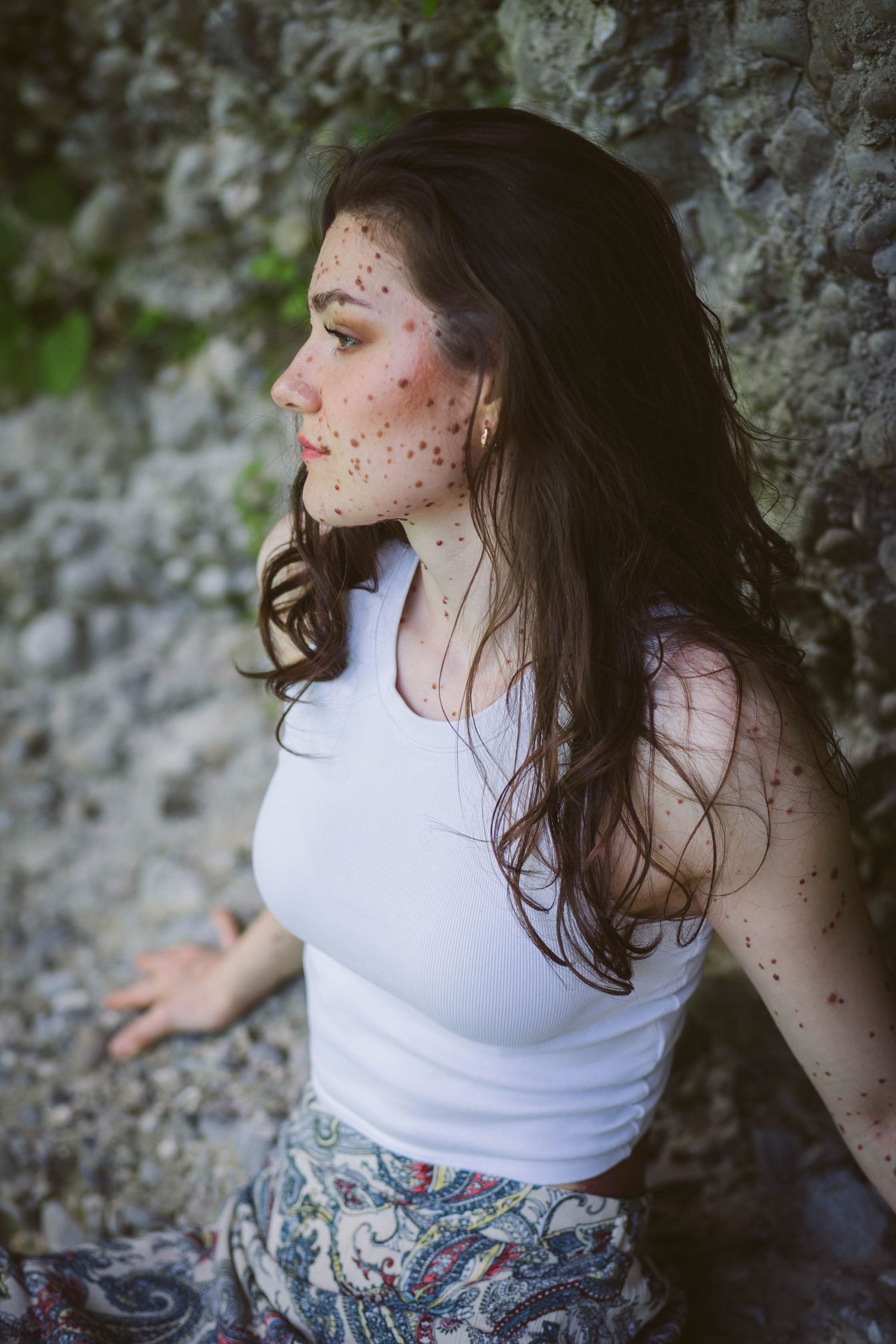 Person wearing white tank top with long dark hair and freckles poses against stone wall in artistic portrait.