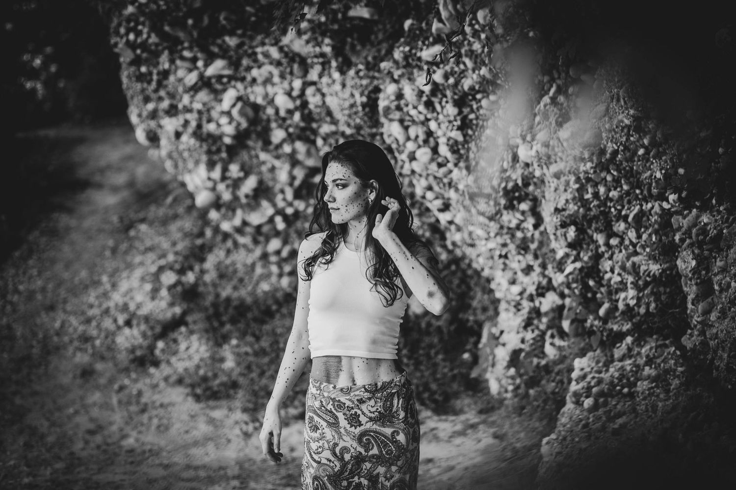 A young woman in a crop top and patterned skirt stands thoughtfully on a garden path in black and white photography.