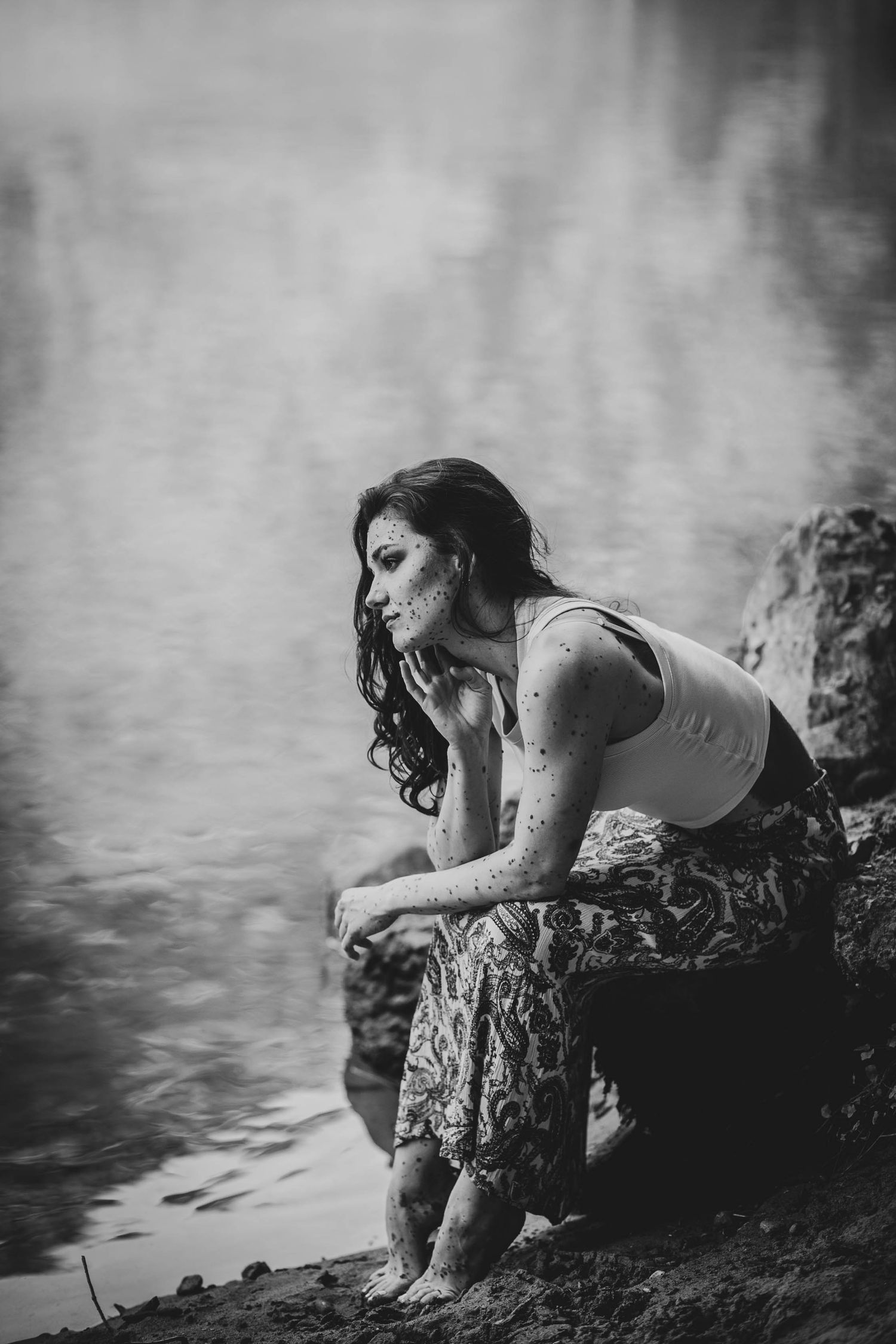 Black and white portrait of a woman in casual attire sitting on rocks near calm water in a reflective moment.