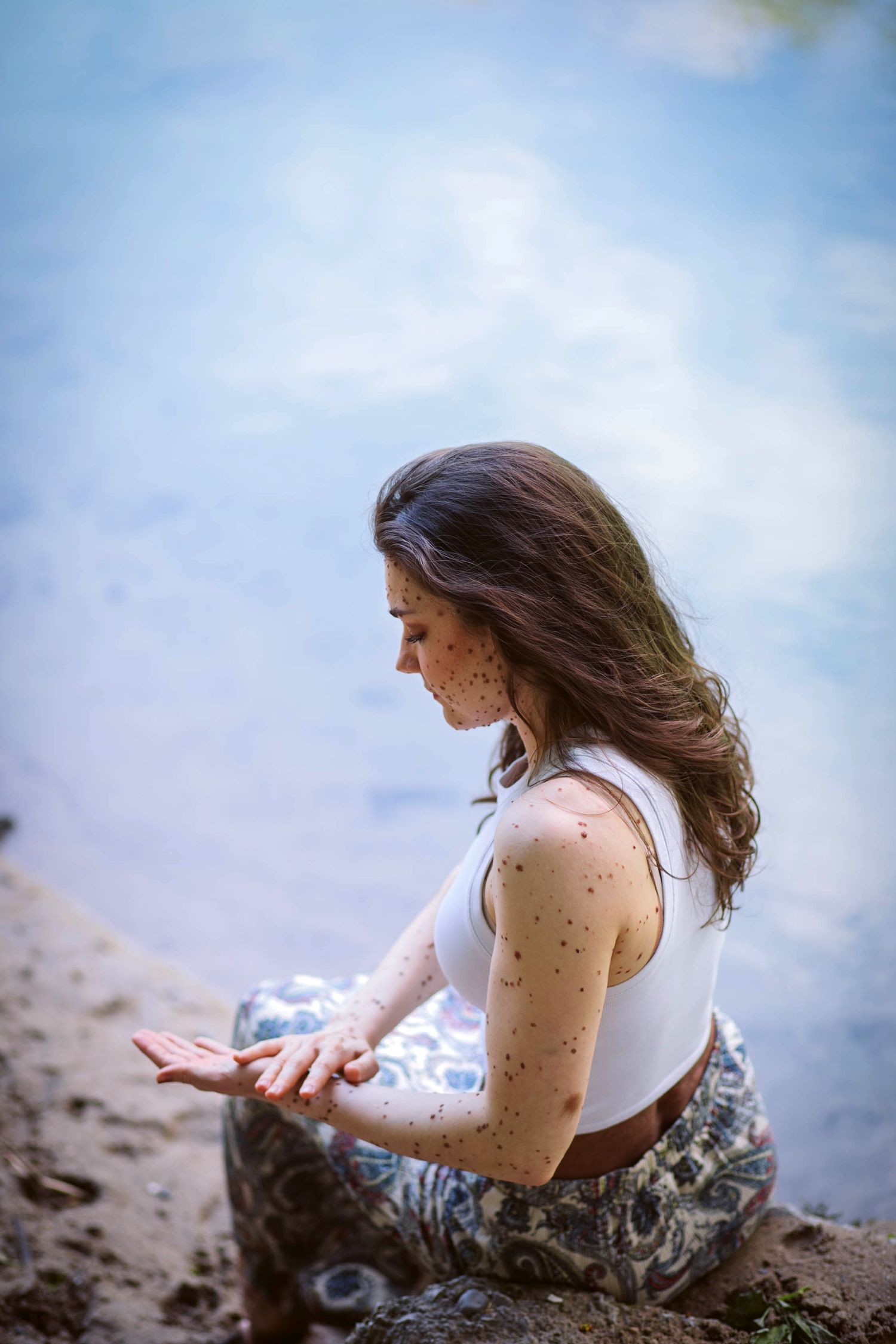 A young person in a white tank top and patterned pants sits by the water's edge on a tranquil day.