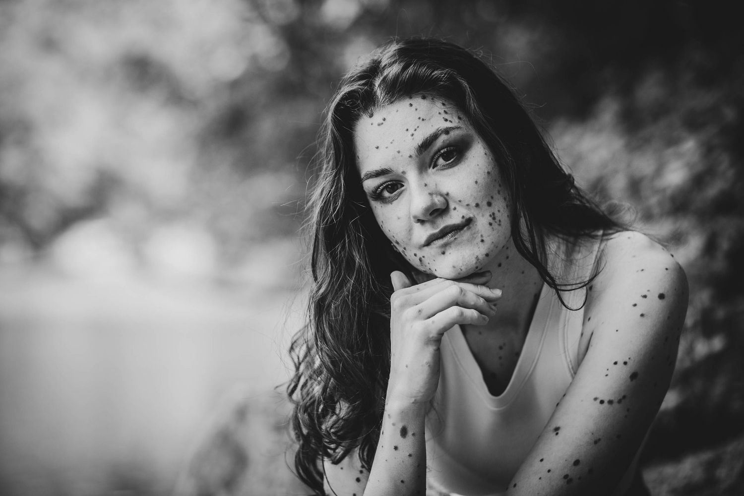 Black and white portrait of someone with long dark hair and delicate freckles sitting in a contemplative pose outdoors.