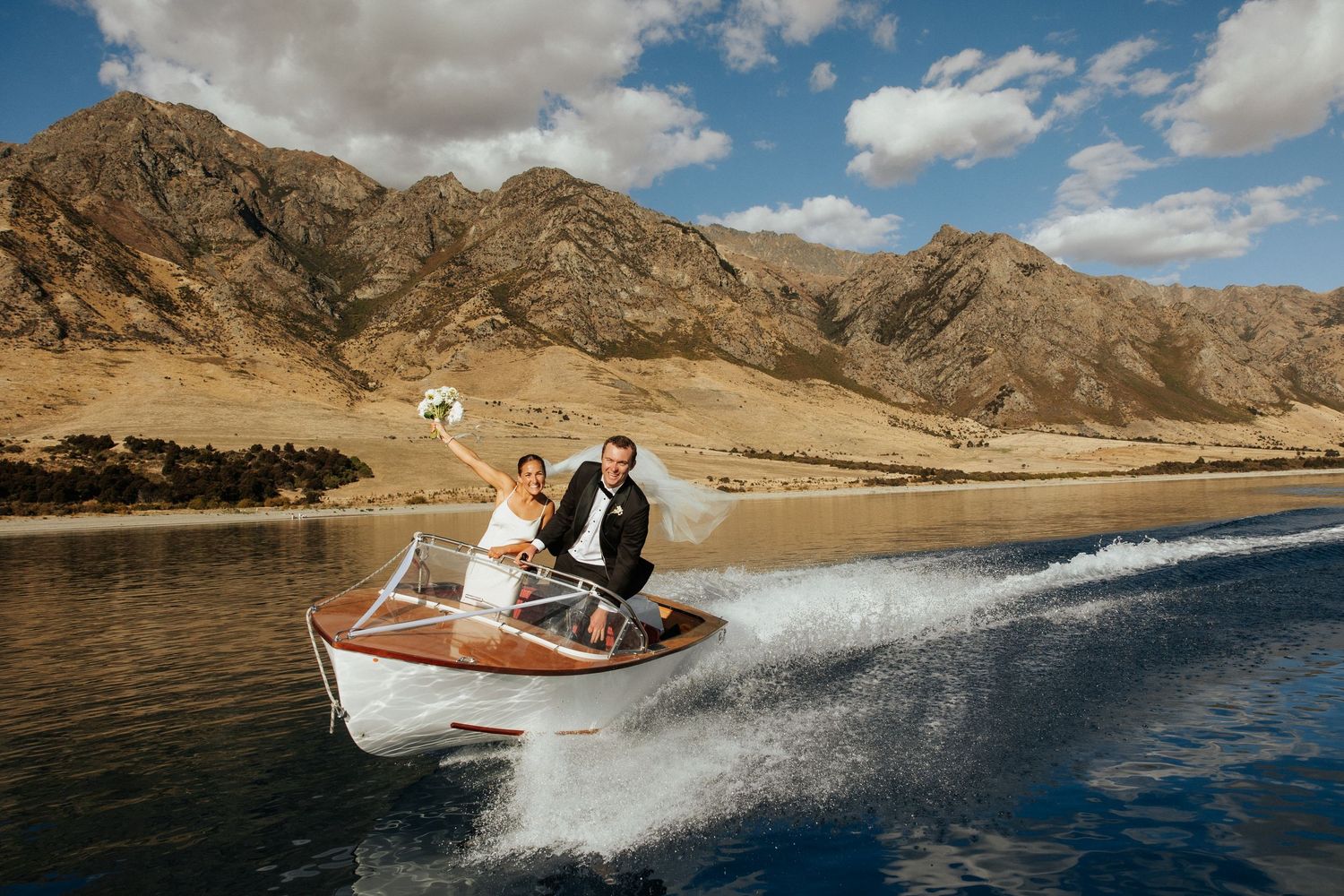 Two people ride in a speedboat across a lake with rugged mountains and blue sky in the background, wanaka