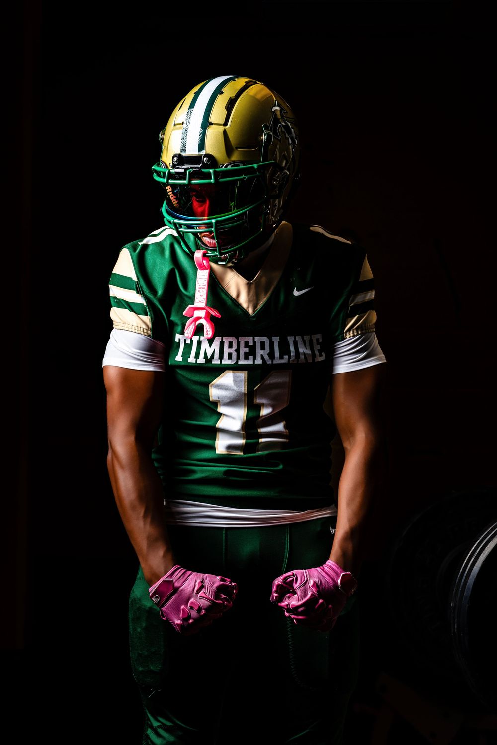 Timberline football player in green and gold uniform with pink gloves poses dramatically against black background.
