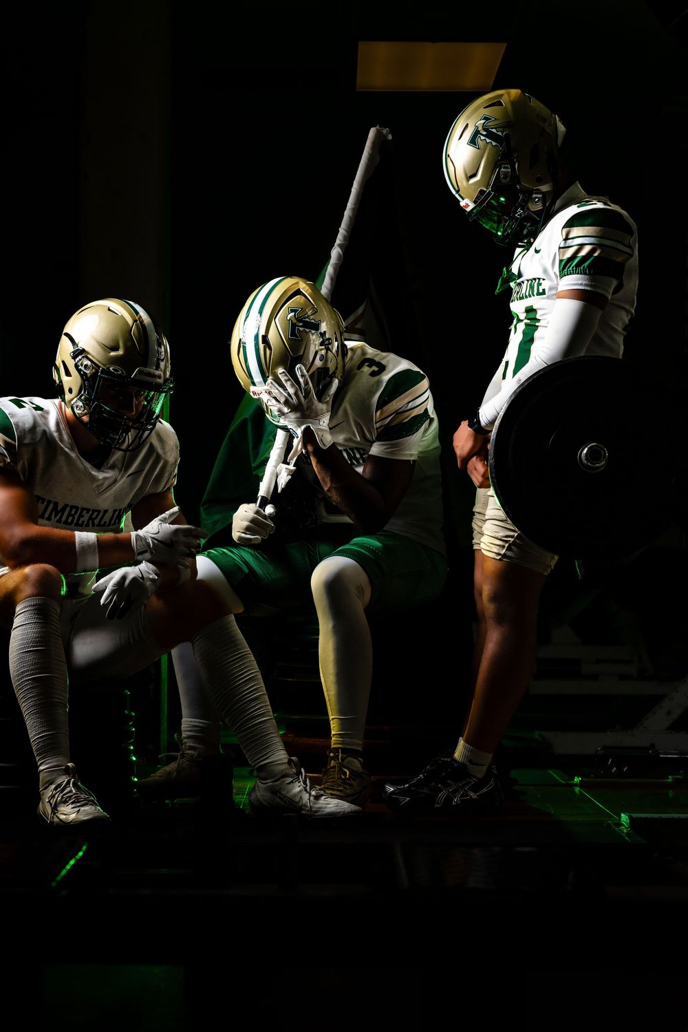 Three football players in green and gold uniforms sit in a dark tunnel, heads bowed in pre-game prayer.