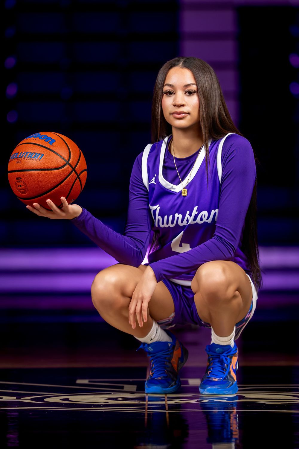 A young female basketball player in a purple Thurston jersey kneels on a court, holding a basketball in a professional sports portrait.