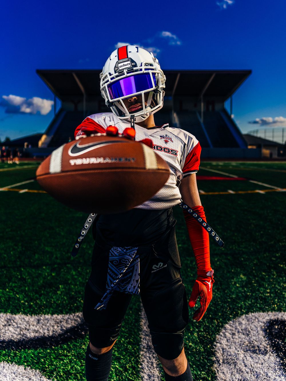 Football player in white helmet and red gloves holds ball toward camera on turf field.