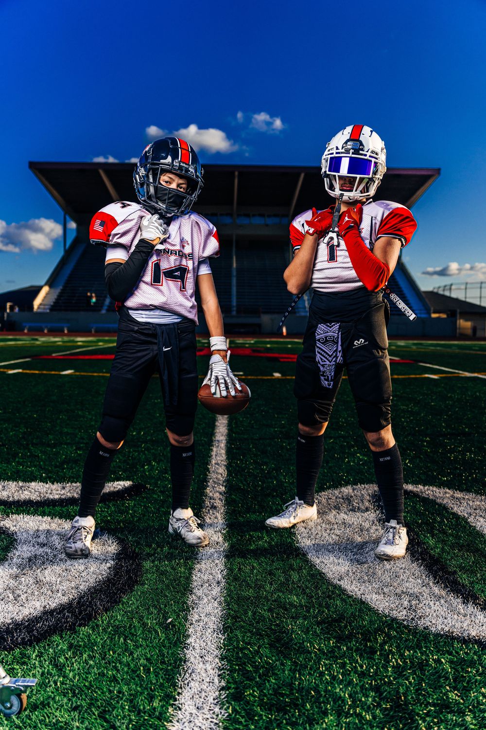 Two youth football players in red and white uniforms pose on a green turf field under a blue sky.