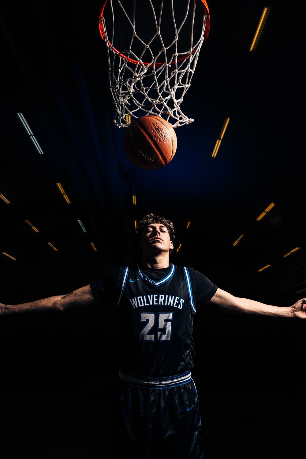 Basketball player #25 in Wolverines jersey tosses ball toward hoop against dramatic dark background.