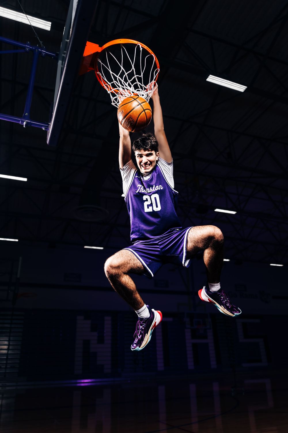 Basketball player #20 in purple jersey dunks ball through hoop mid-air in indoor gym.