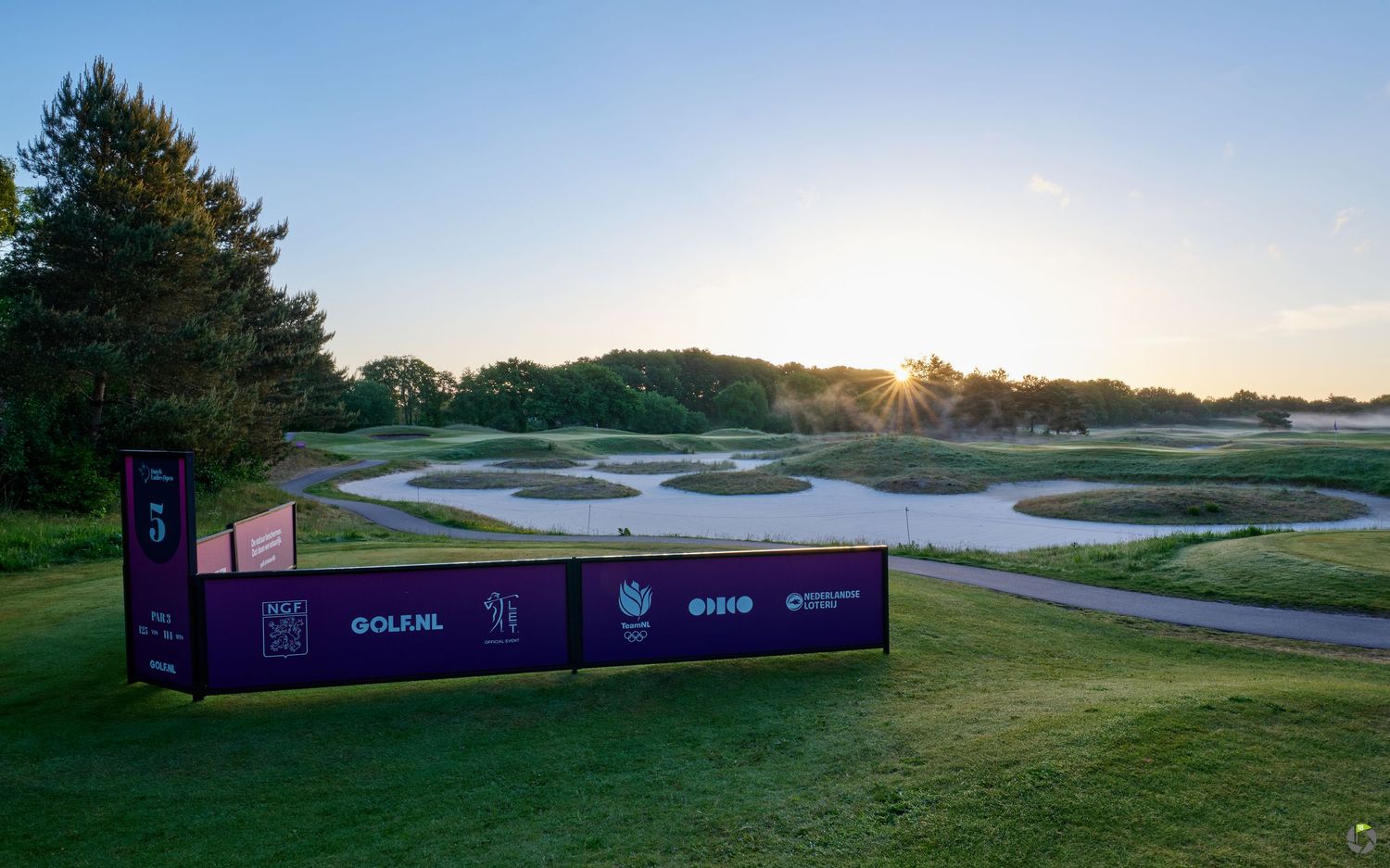 A purple tournament banner sits beside a golf course fairway with sand bunkers at sunrise.