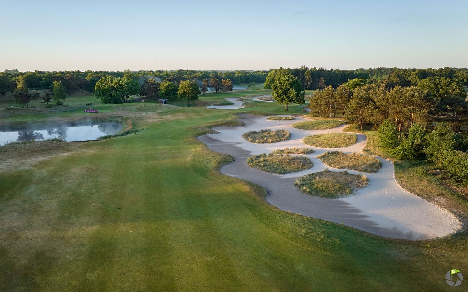 Aerial panoramic view of a golf course showcasing winding paths, sand bunkers and lush green fairways surrounded by trees.
