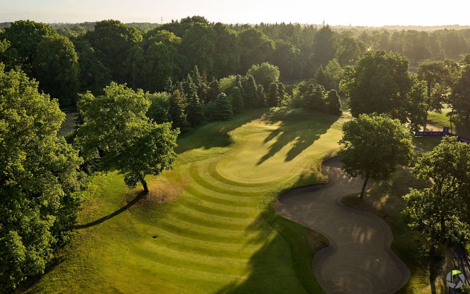 Aerial view of a golf course fairway with curved stripes and surrounding trees at golden hour.