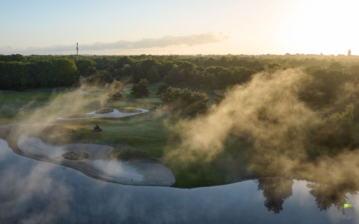 Morning mist rises over a scenic golf course with winding waterways and lush greenery at sunrise.