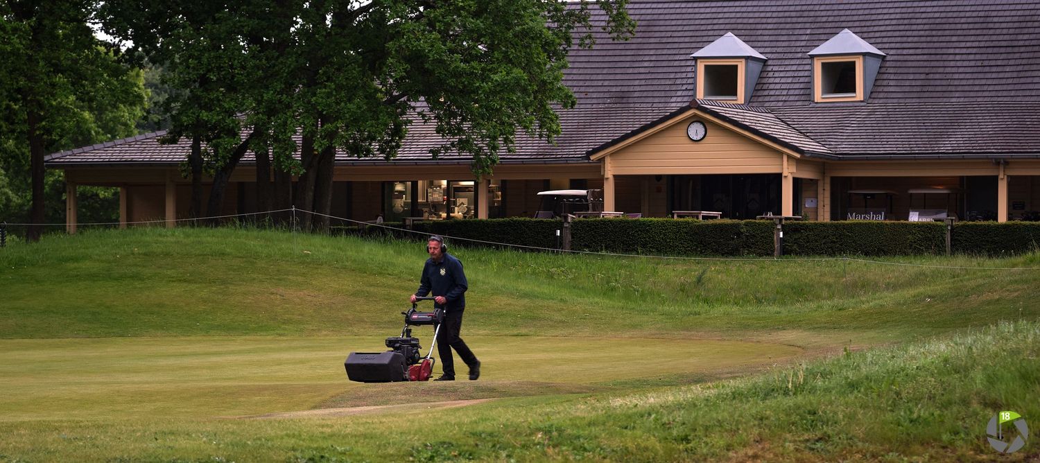 Groundskeeper mowing grass on a golf course with residential buildings in the background.