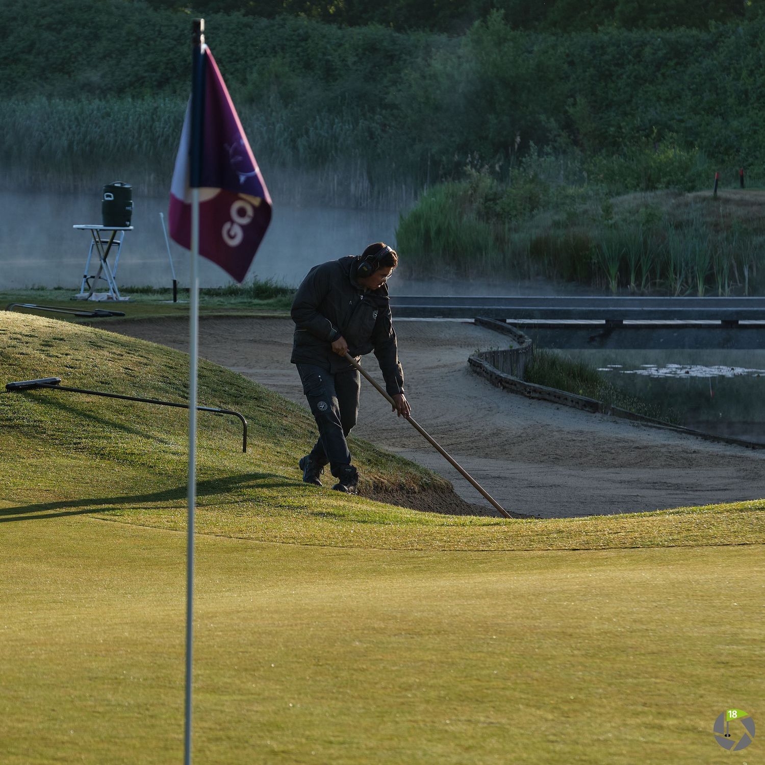 Maintenance worker raking sand near a golf course flag during early morning hours.