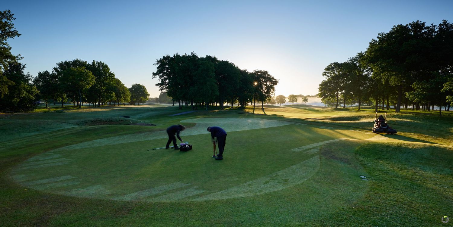 Two golfers practice putting on a lush green golf course at sunrise with trees silhouetted against the sky.