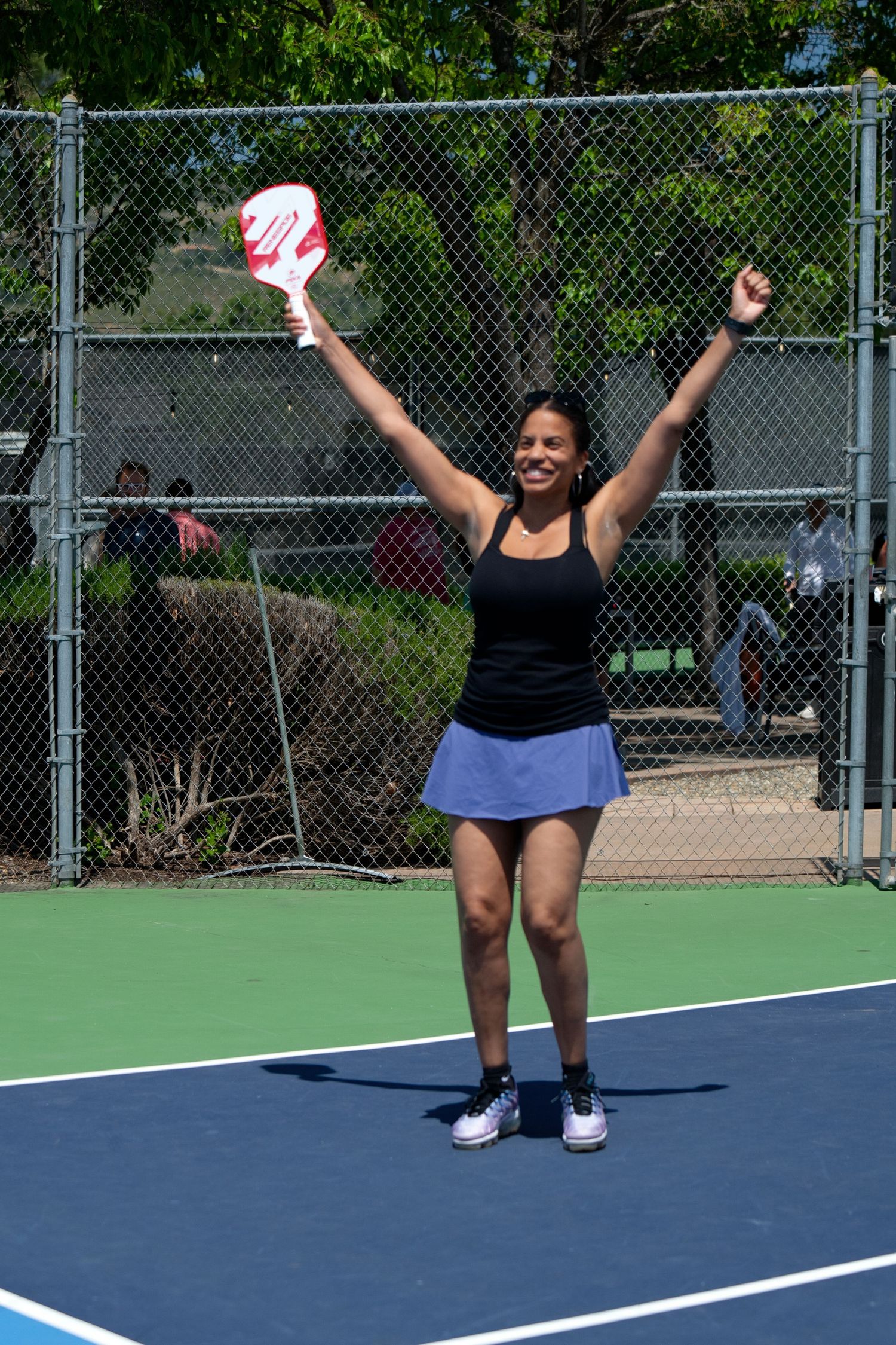 Athlete celebrates with raised arms on outdoor tennis court holding pickleball paddle and ball.