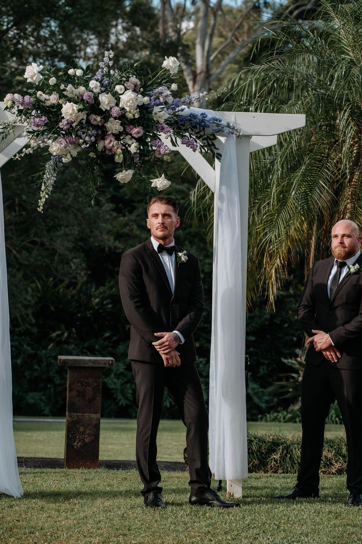 Wedding ceremony setup with a white pergola decorated with pink and white floral arrangements on a green lawn.