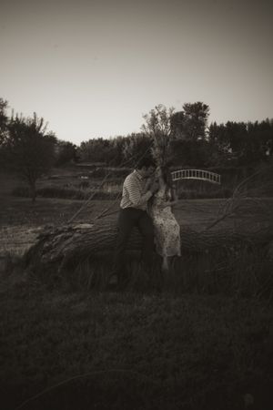 A vintage-style photo of a couple embracing near a bridge in a rural setting.
