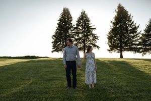 Romantic couple standing in a grassy field at sunset with tall evergreen trees in the background.