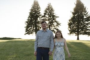 A couple in coordinated outfits standing together in front of tall pine trees at sunset.
