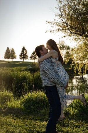 Couple in romantic embrace against sunset backdrop with pond and trees.
