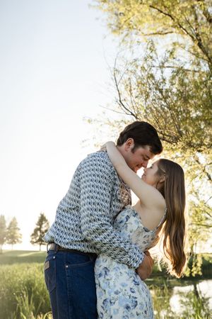 Romantic couple embracing in meadow with water and trees in background.