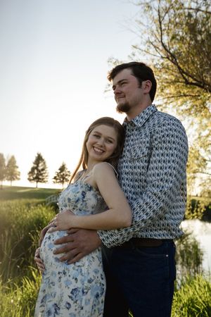 Happy couple embracing in sunlit field with pond and trees in background.