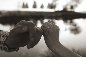 Close-up of hands exchanging an engagement ring by a lake at sunset.