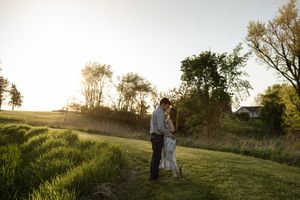 A couple stands in a sunlit field during golden hour with tall grass and trees in the background.
