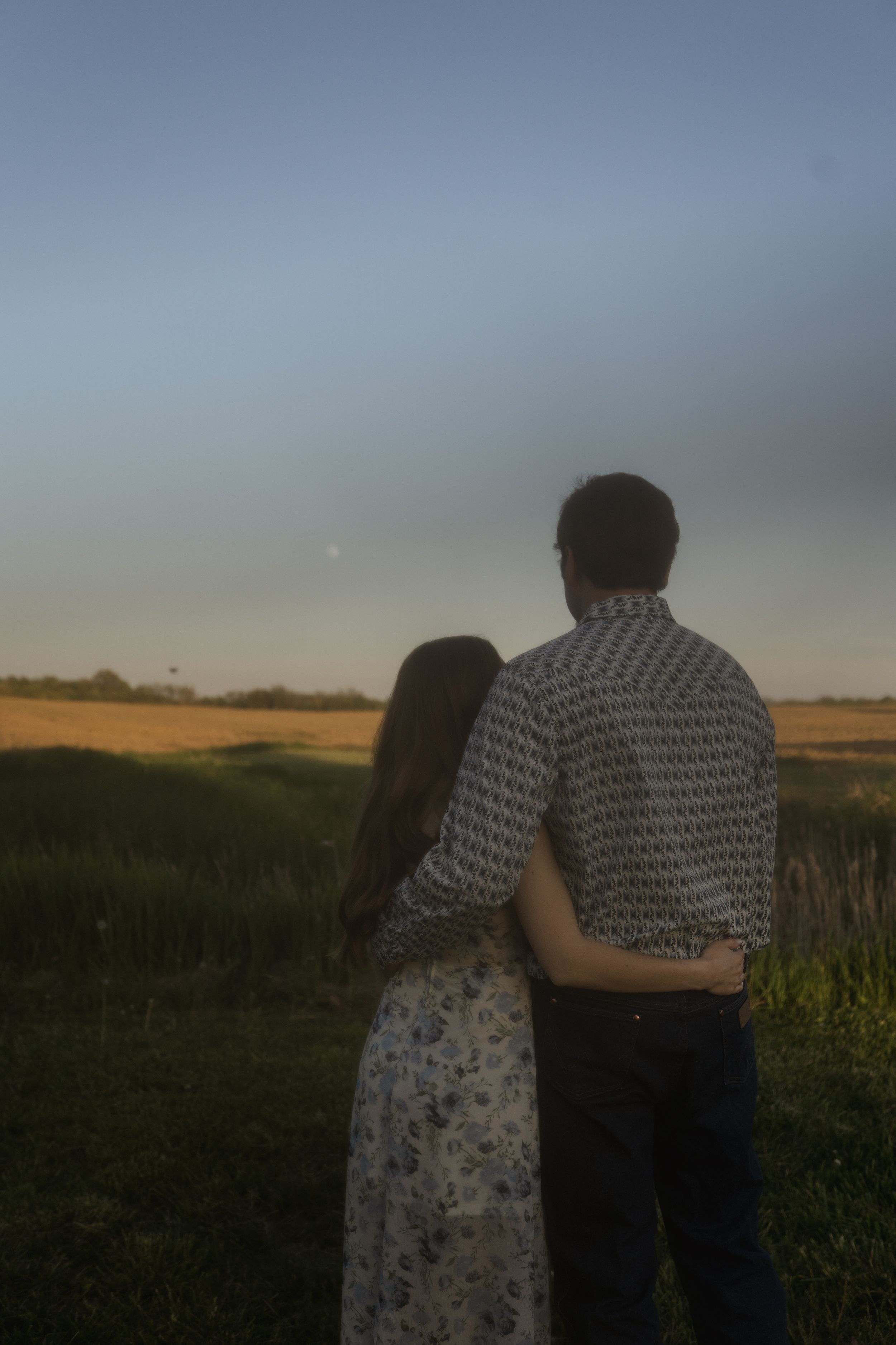 Couple embraces while standing in a golden field during sunset.