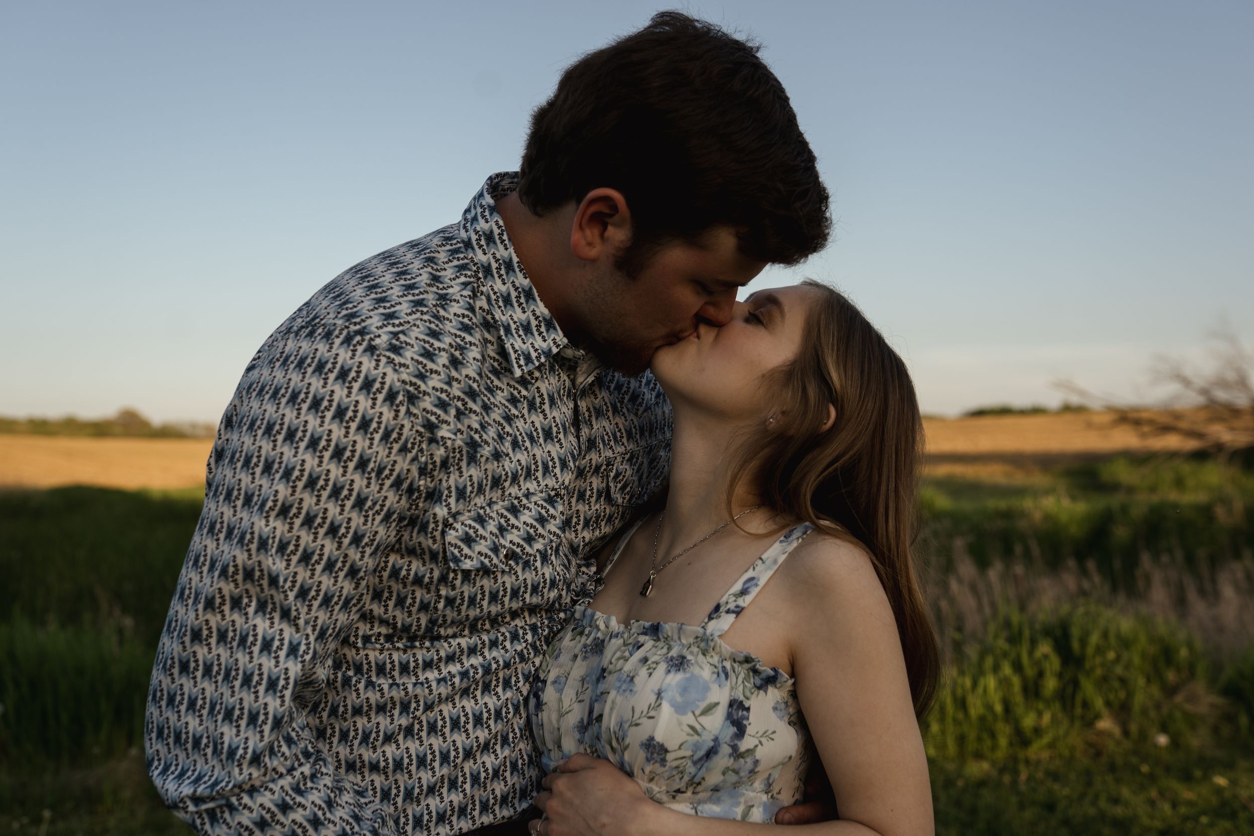 Couple embraces lovingly against sunset backdrop in rural setting.