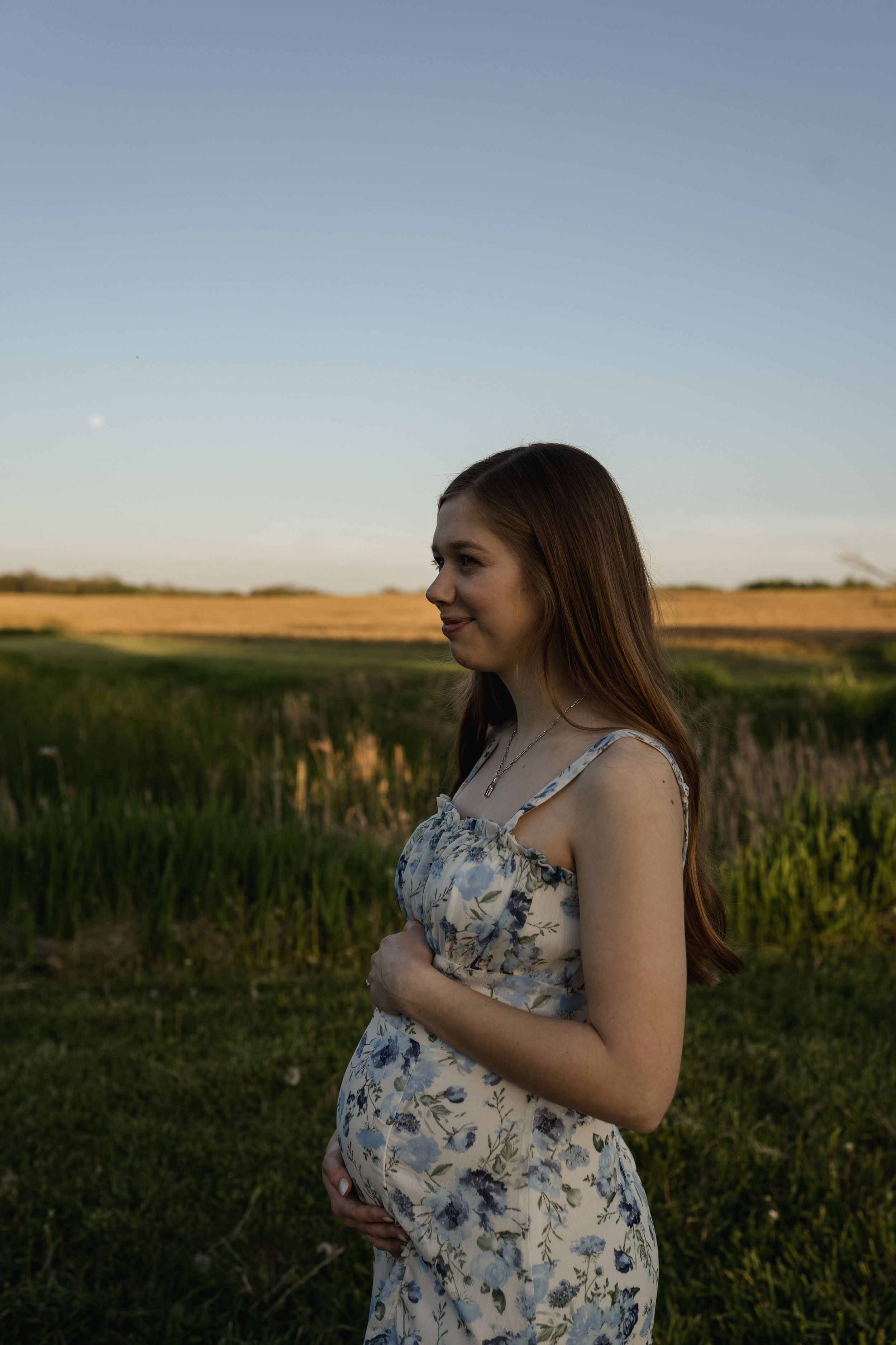A floral dress silhouette stands against a rural sunset landscape with fields in the background.