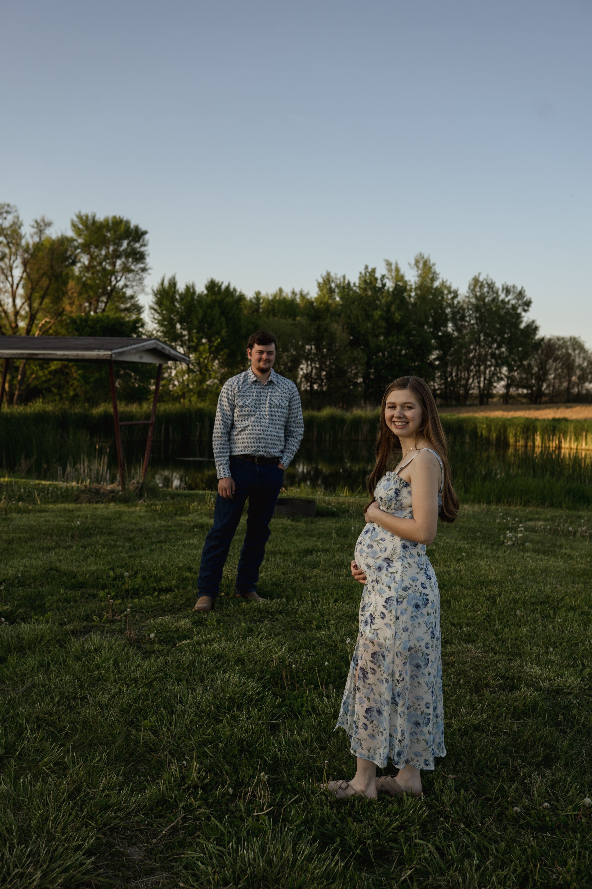 A couple stands apart in a grassy field during golden hour with trees in the distance.