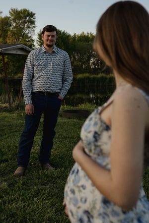 A couple stands in a grassy meadow during sunset with one wearing a patterned shirt and jeans.