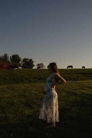 A person in a floral dress stands in a field at dusk with horses visible on the horizon.