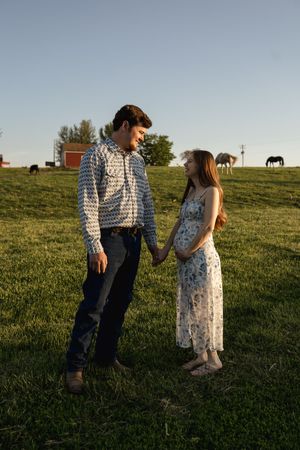 A couple shares a tender moment in a sunlit field with horses grazing in the background.