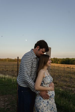 Couple in patterned shirt and floral dress embrace near farm fence at dusk.