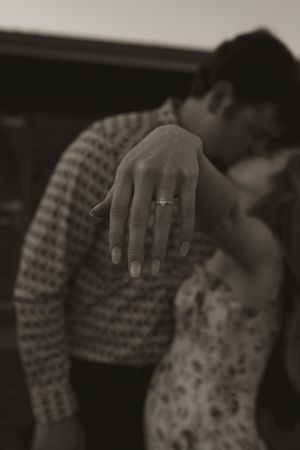 A close-up black and white photo showing a hand with an engagement ring against a blurred background.