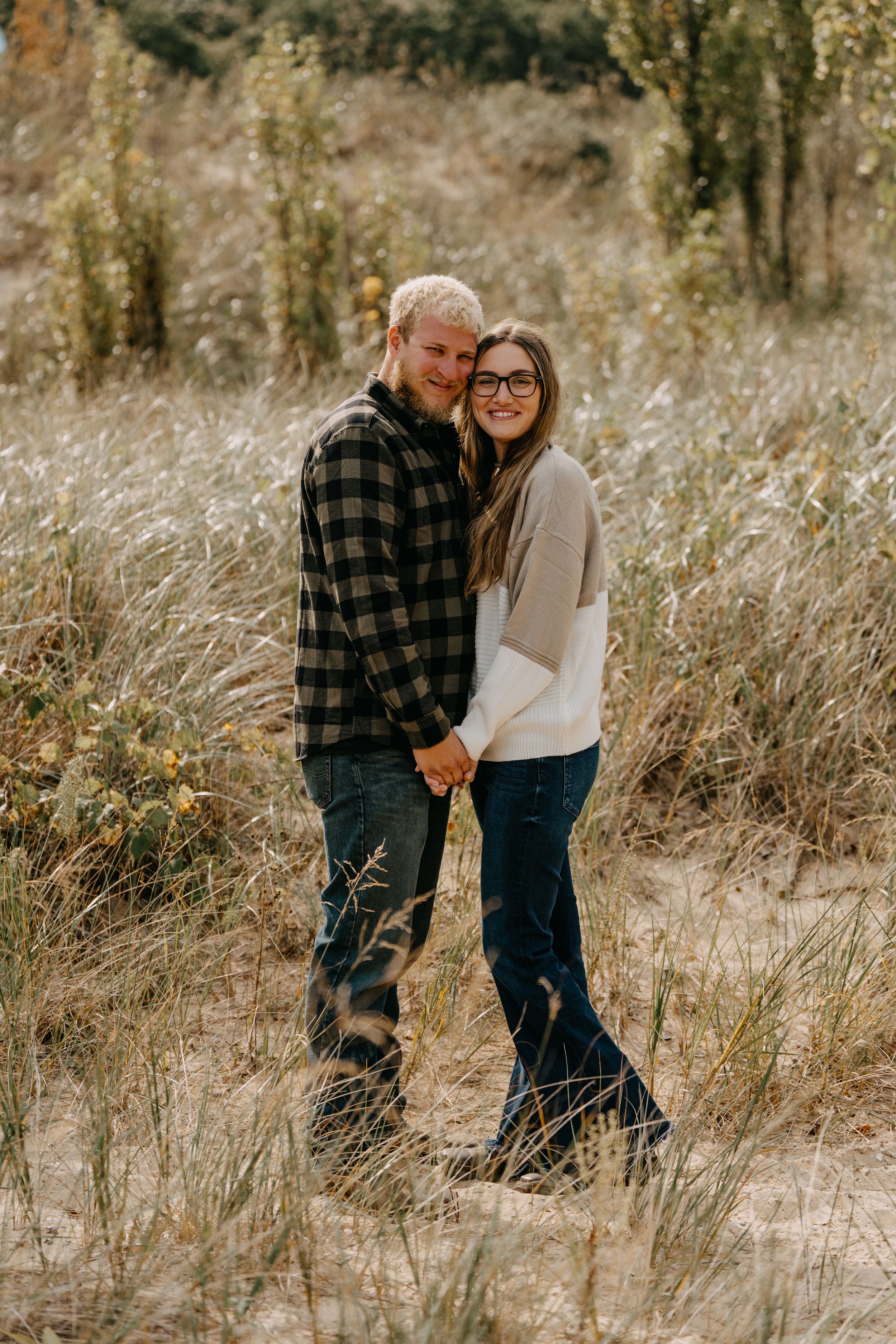 A couple stands together in tall golden grass on an autumn day wearing casual clothing and holding hands.
