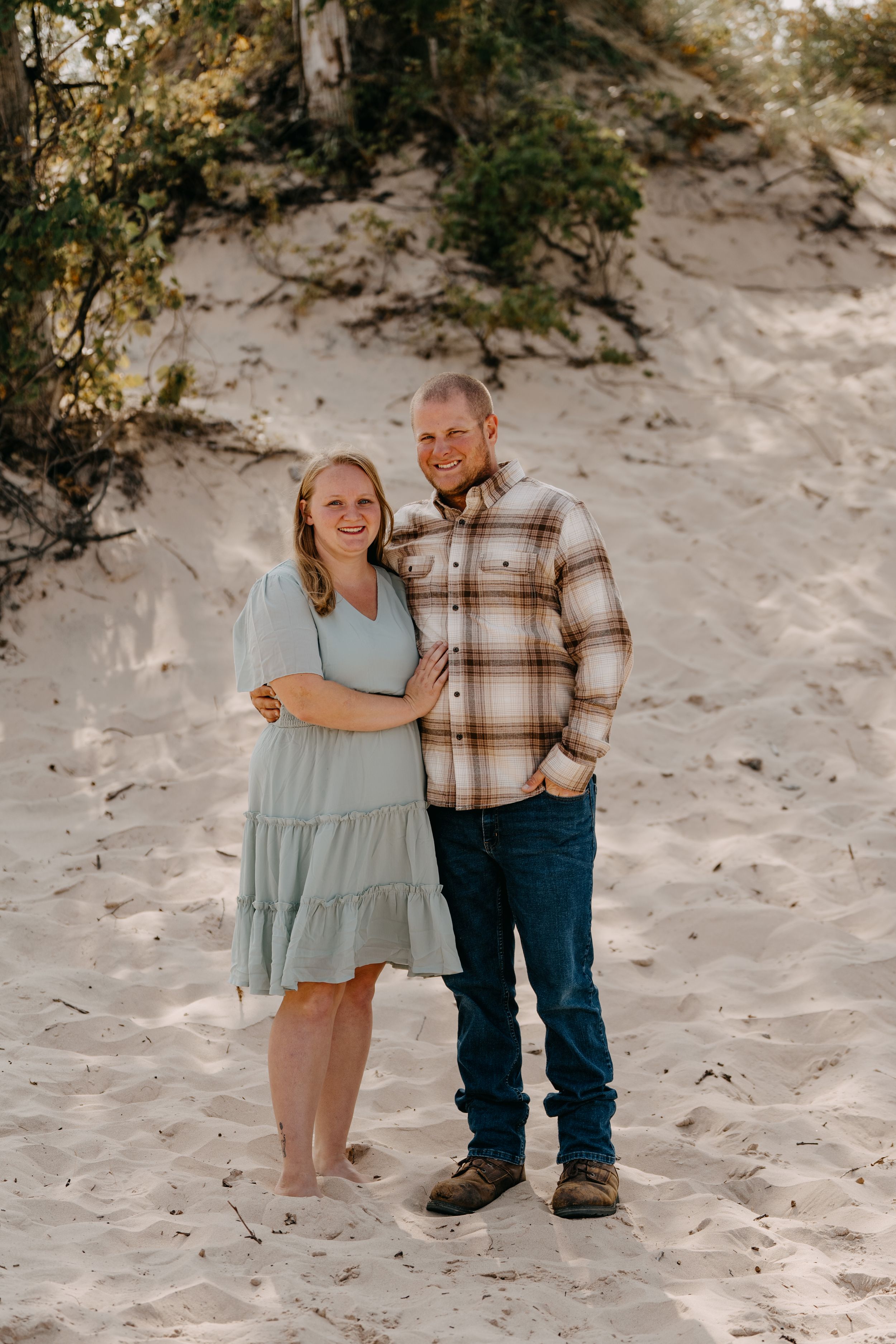 A couple walks hand in hand along a sandy beach path surrounded by coastal vegetation on a sunny day.