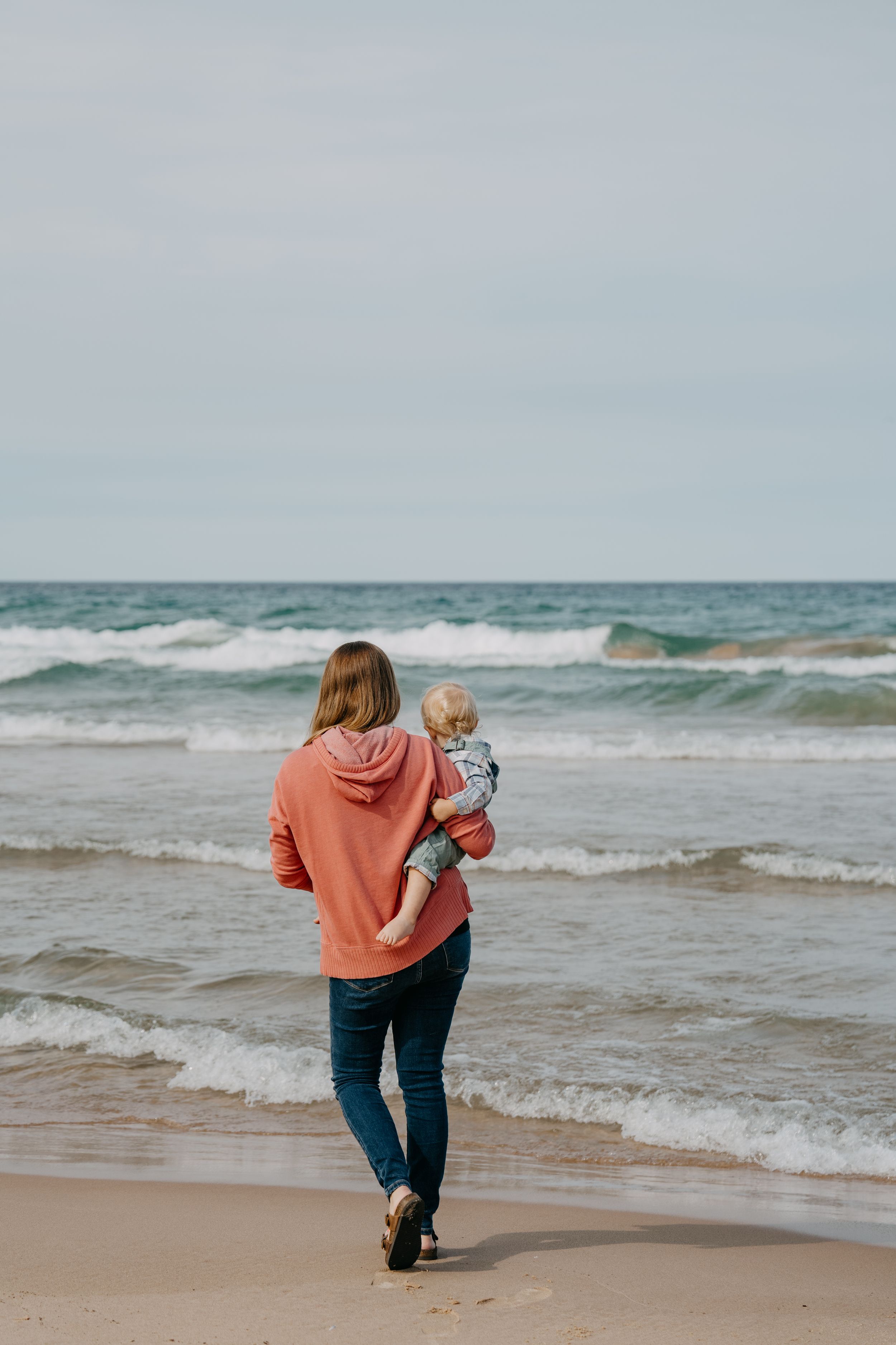 Person in coral hoodie holds small child while walking along sandy beach with crashing waves on overcast day.