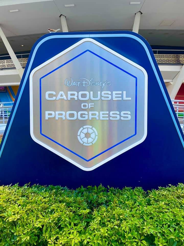Disney theme park signs featuring Carousel of Progress and Progress logos in blue hexagonal shapes above green hedges.