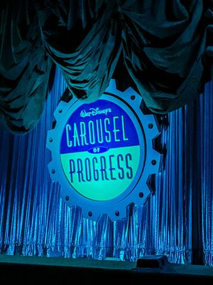 Illuminated blue Carousel of Progress sign glows against dark curtains at Walt Disney's theme park attraction.