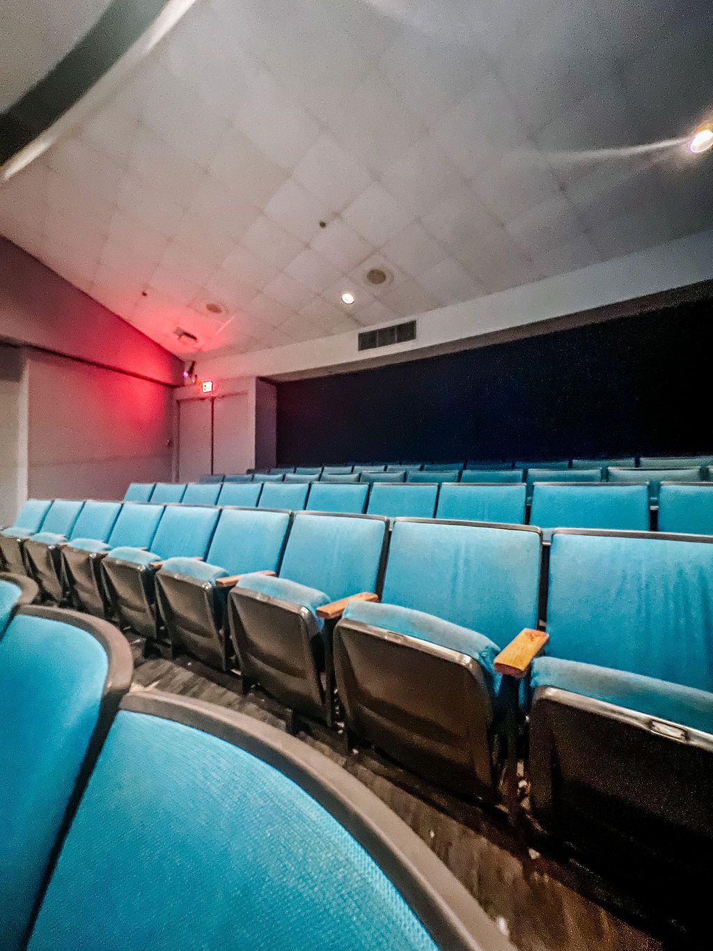Empty theater auditorium with curved rows of bright turquoise seats illuminated by red and white lighting.