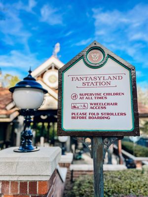 A Fantasyland Station sign at Disney theme park with instructions for guests and a decorative lamp post against blue sky.